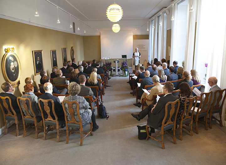 Perspektiv från längst bak i sal med publikrader, i fonden högtidligt instiftande av Stiftelsen för Sveriges unga akademi, Anna Sjöström Douagi, dåvarande verksamhetschef.